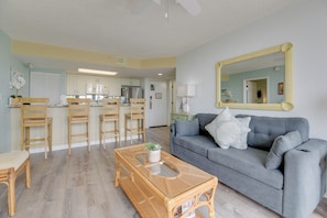 A living room with a grey sofa, wooden chairs, a glass coffee table, and a kitchen in the background. - The Samana Cay | Tropical, Airy Retreat w/ Private Balcony, Pool! (Key West)