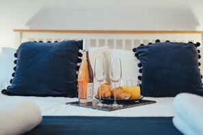 Un petit-déjeuner soigneusement préparé est disposé sur un lit agrémenté d’oreillers bleu foncé, avec une bouteille de jus d’orange, deux verres et des croissants.