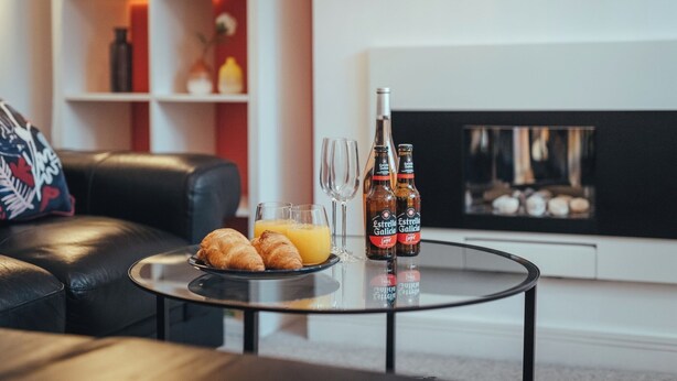 Une table en verre sur laquelle sont disposés des croissants, du jus d’orange et deux bouteilles de bière Estrella Galicia, le tout dans un salon moderne doté d’une cheminée.