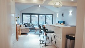 A modern kitchen with a dining area, featuring a bar stool set, a sofa, and a large window offering a view of the city. - Contractors Retreat- Free Parking (Chelmsford)