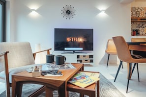 Una sala de estar moderna con un televisor de pantalla plana mostrando 'NETFLIX', una mesa de centro de madera con revistas, una lámpara y una silla.