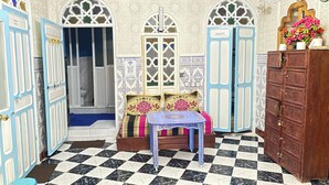 A room with a checkered floor, a bed with patterned pillows, a small table, and a wooden cabinet with numbered drawers. - aline hostel (Chefchaouen)