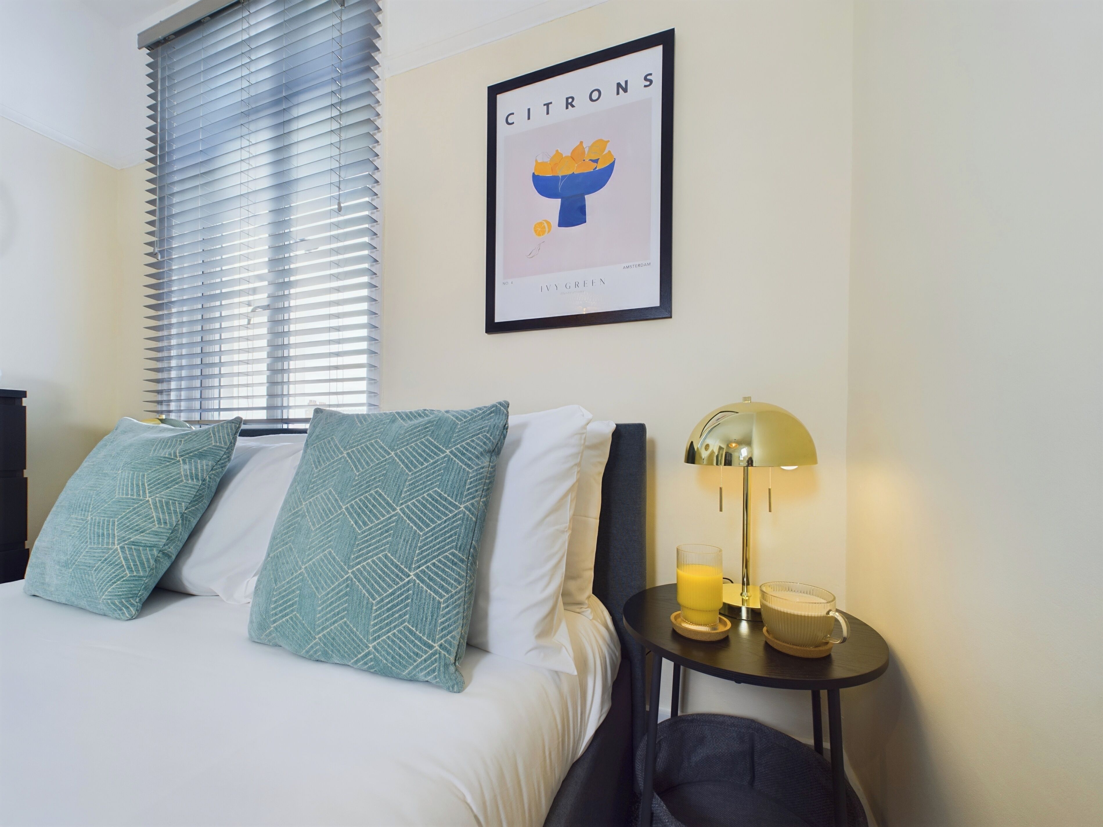 Habitación de hotel con cama, dos almohadas estampadas, mesita de noche con lámpara, un cuadro enmarcado en la pared y una ventana con persianas.