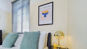 A hotel room with a bed, two patterned pillows, a nightstand with a lamp, a framed picture on the wall, and a window with blinds. - Seaside Haven Retreat in Southend (Southend-on-Sea)