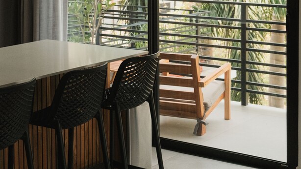 Ein moderner Essbereich mit einem weißen Tisch und schwarzen Stühlen, einer gewebten Pendelleuchte und Blick auf einen Balkon mit Geländer und Palmen.