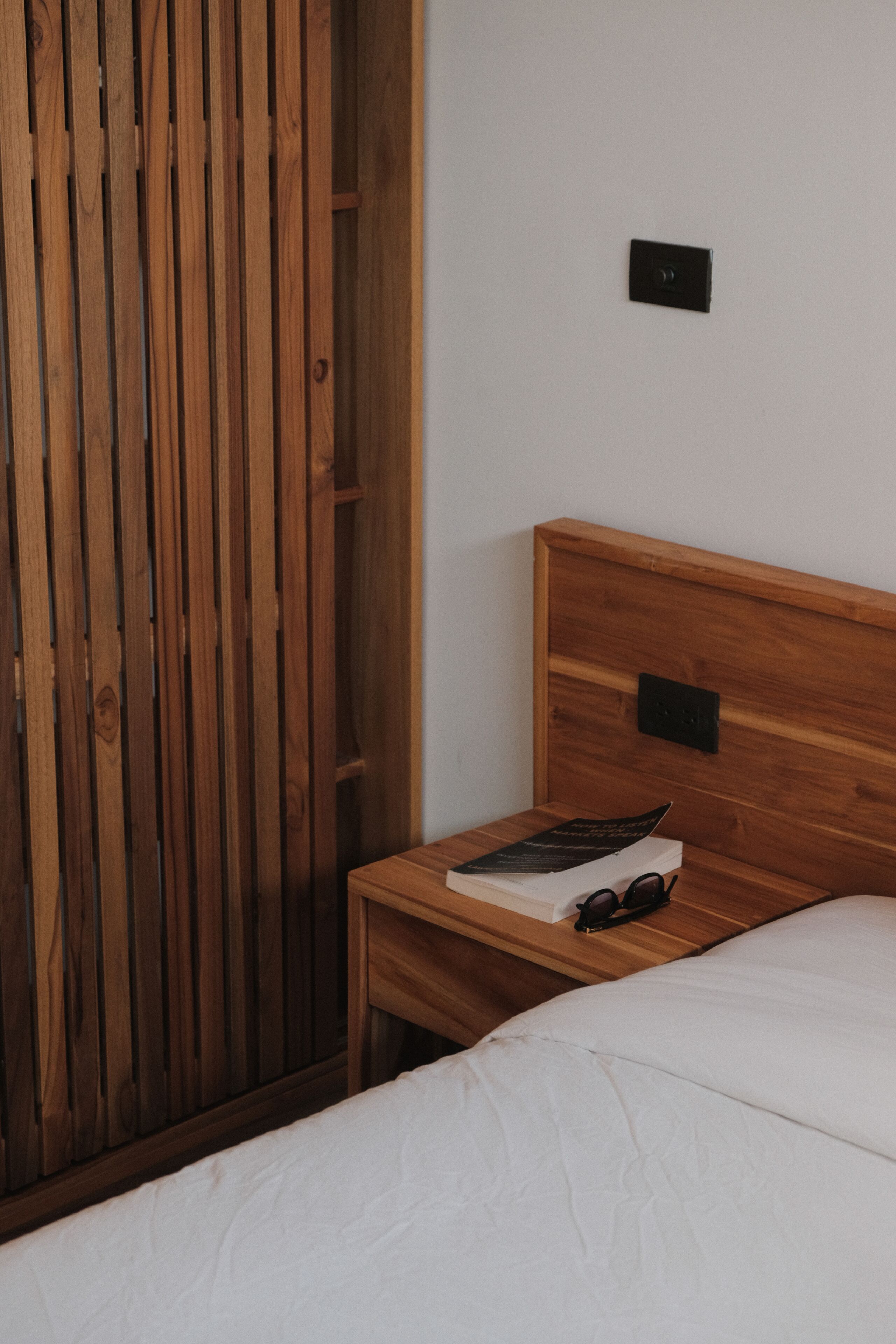 A wooden bed with a white mattress, a wooden nightstand with a book and glasses, and a wooden wardrobe.