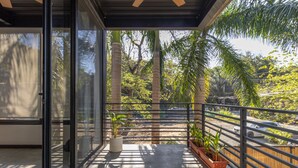 Un balcon doté d’une porte vitrée, de plantes en pot et offrant une vue sur des palmiers et une rue.