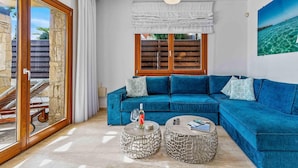Un salon avec un canapé d’angle bleu, deux petites tables basses rondes en pierre, une fenêtre avec des volets en bois et une vue sur la mer.