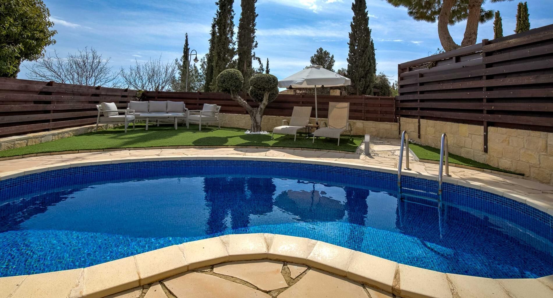 A swimming pool with a tiled edge, surrounded by a wooden fence, and a grassy area with lounge chairs and a tree.