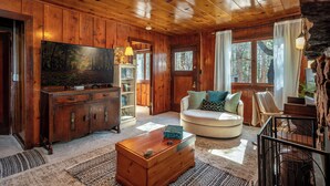 A cozy cabin interior with wooden walls, a large TV, a round sofa, and a wooden coffee table.
