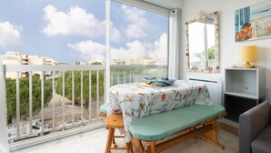 A balcony with a table and bench, a large window with a view of buildings and trees, and a mirror on the wall. - Cozy Home by the Sea (Le Grau-du-Roi)