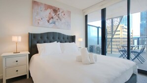 Un grand lit avec du linge de lit blanc, une tête de lit capitonnée et deux tables de chevet avec des lampes. Il y a une grande fenêtre donnant sur des immeubles et un balcon avec des chaises.