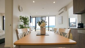 Un espace de repas moderne avec une table en bois et des chaises blanches, une plante en pot, et une vue sur la ville grâce à de grandes fenêtres.