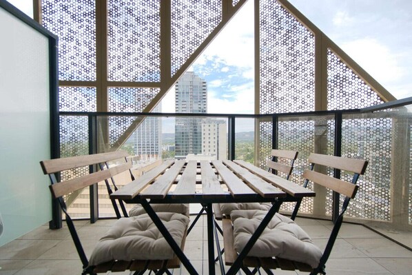 A balcony with a wooden table and benches, large windows with a geometric pattern, and a view of city buildings. - Beautiful & Comfortable 2BR Apt in Rundle Mall (Adelaide)
