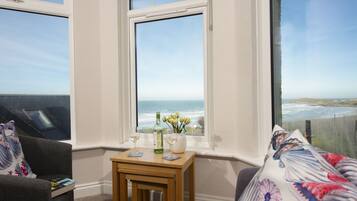 A room with a view of the ocean, a small wooden table with a bottle and glass, and a couch with floral cushions.