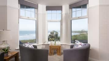 A corner room with three large windows overlooking the ocean, a small table with a vase of flowers, and two grey armchairs.