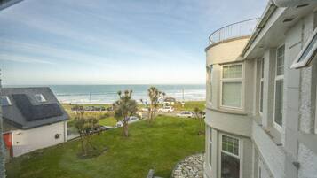 A coastal view from a balcony overlooking a grassy area with parked cars, a beach, and a clear sky.