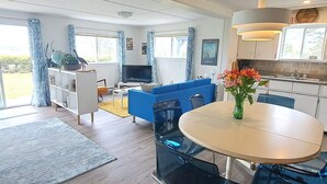 A modern living room with a blue sofa, a dining table with chairs, and a kitchen area with a sink and cabinets. - Bright, Spacious Beachwalk Cottage: Ocean view, 5 minutes to sandy beaches (Sable River)