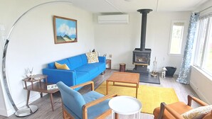 A living room with a blue sofa, wooden chairs, a small table, a yellow rug, a fireplace, and a painting on the wall. - Bright, Spacious Beachwalk Cottage: Ocean view, 5 minutes to sandy beaches (Sable River)