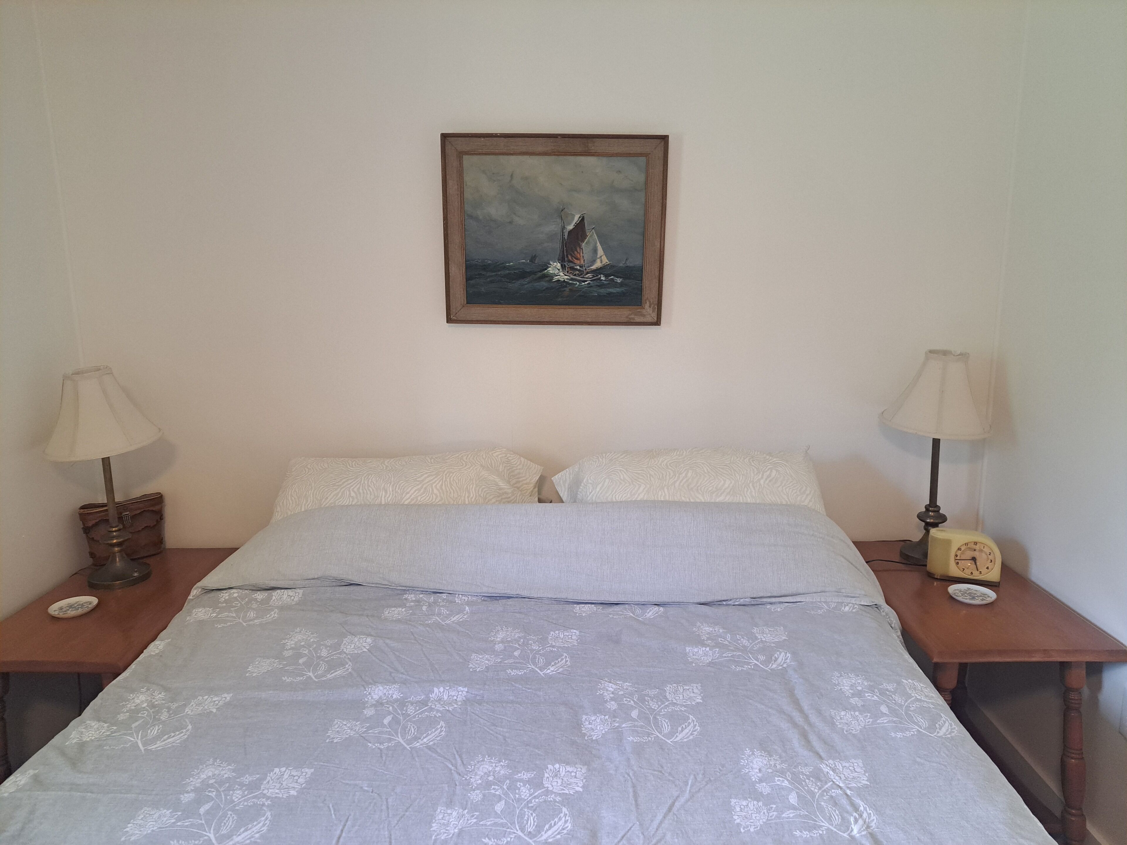 A bed with floral-patterned bedding, two bedside tables with lamps, a framed painting of a sailboat, and a small clock on one table.