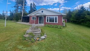 A single-story house with a red exterior and white trim, a front porch, and a small deck. - Bright, Spacious Beachwalk Cottage: Ocean view, 5 minutes to sandy beaches (Sable River)