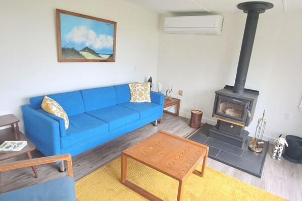 A living room with a blue sofa, a wooden coffee table, a fireplace, and a painting on the wall. - Bright, Spacious Beachwalk Cottage: Ocean view, 5 minutes to sandy beaches (Sable River)