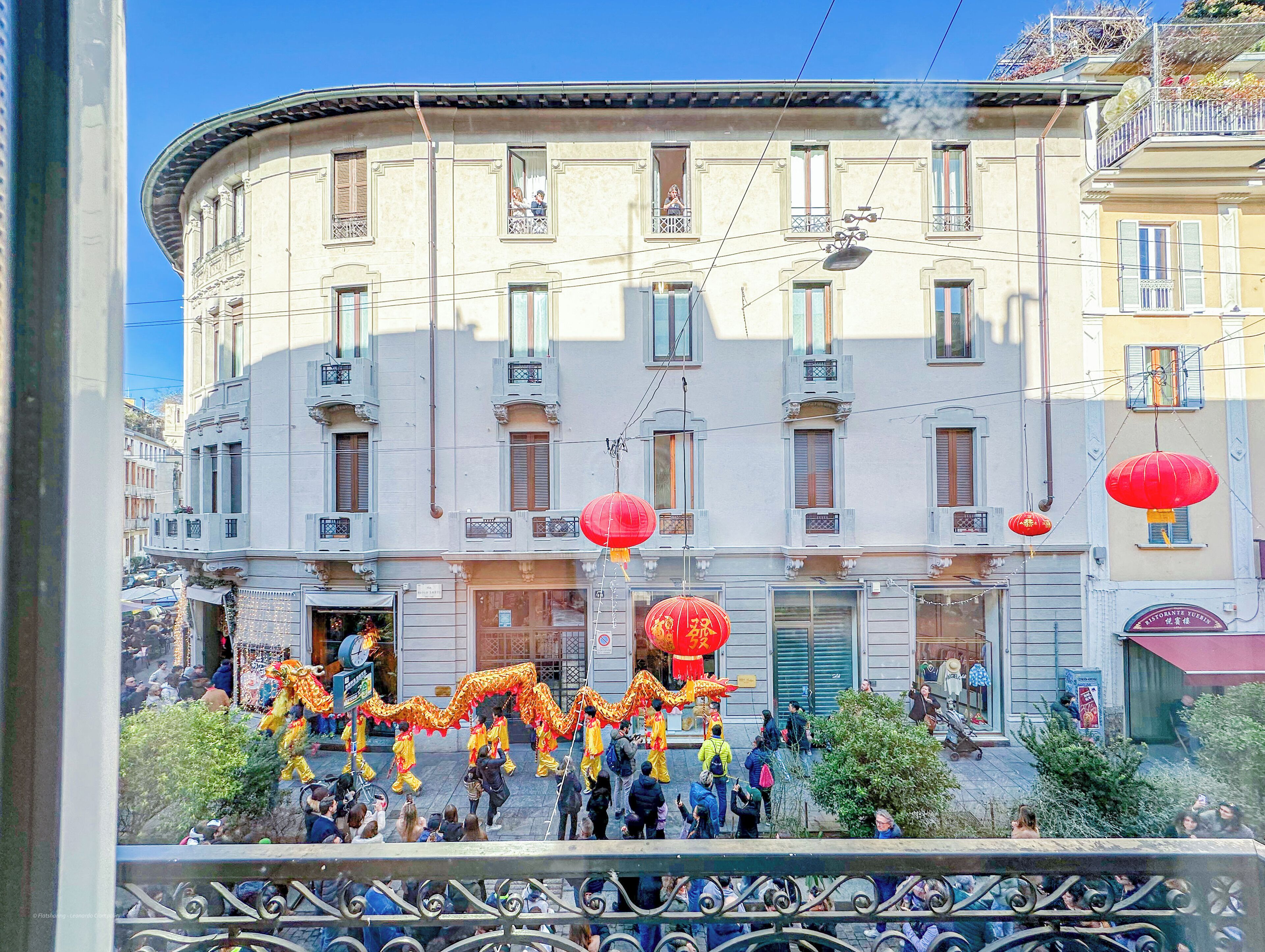Un edificio con varias ventanas y balcones, una calle con peatones y faroles rojos.
