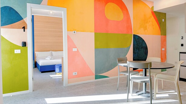 A modern hotel room with a bed, a desk, and a chair. There is a colorful abstract wall mural and a door leading to another room.