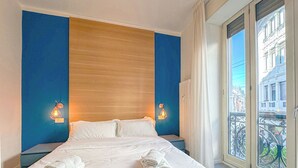 A hotel room with a large bed, two wall-mounted lights, a view of the city through a balcony with iron railings.