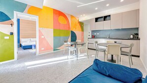 A modern hotel room with a colorful abstract wall, a bed, a blue sofa, a dining area with chairs, and a kitchen with a sink and countertop.