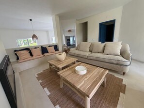 A modern living room with a sofa, two wooden tables, a hanging light fixture, and a view of the kitchen.