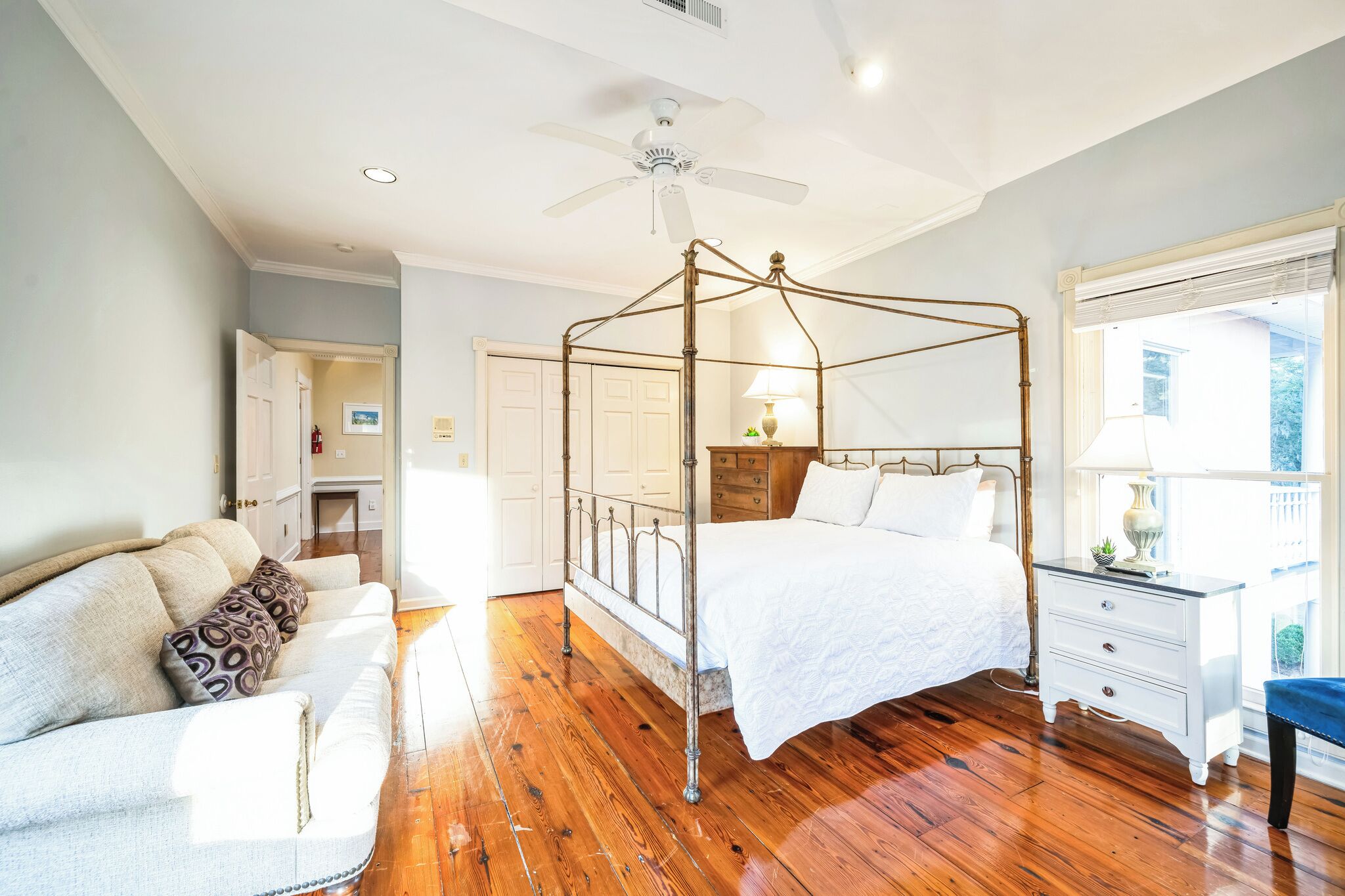 A bedroom with a four-poster bed, wooden flooring, a ceiling fan, and a nightstand with a lamp.