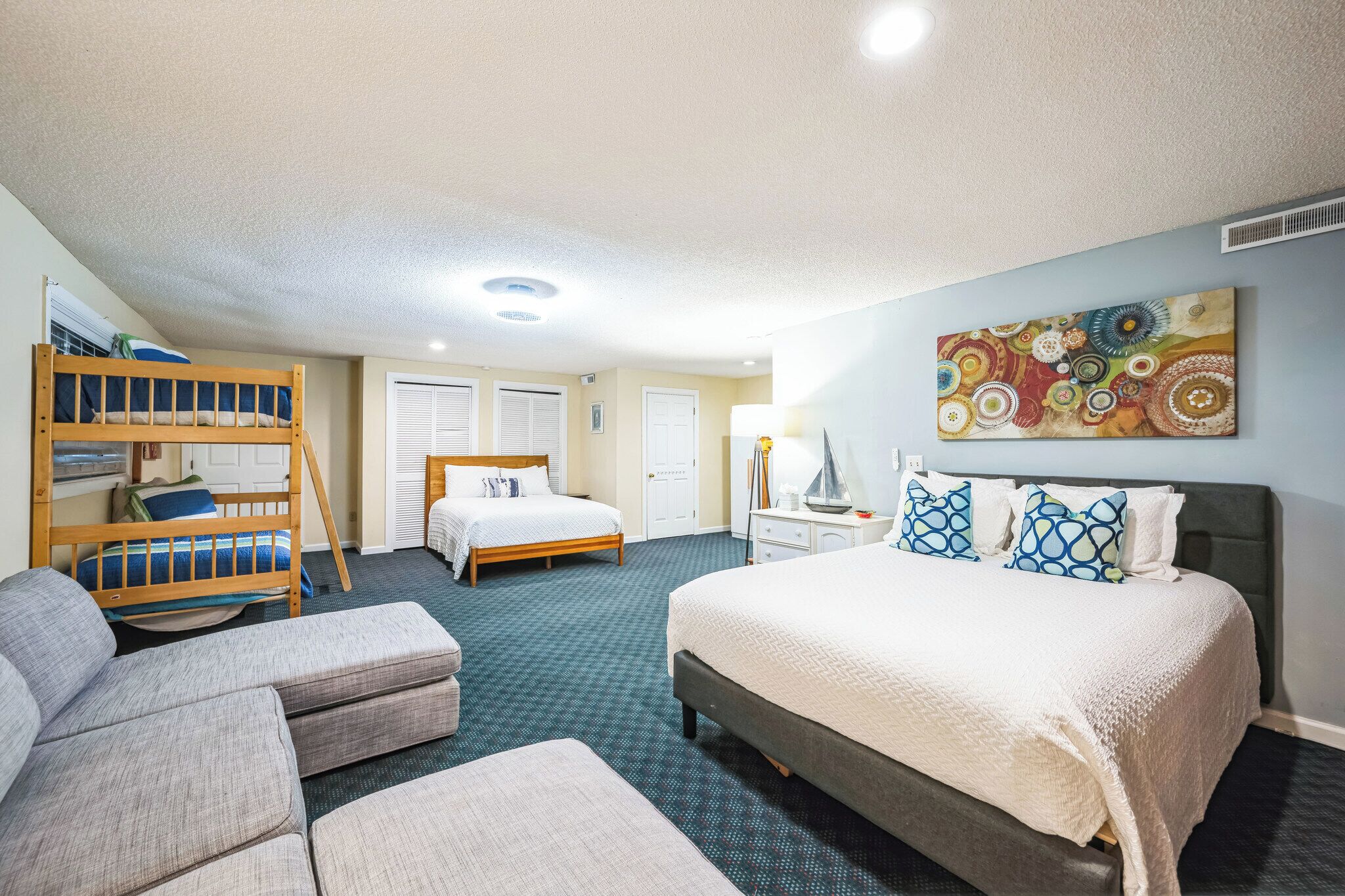 A hotel room with a bunk bed, a single bed, a sofa, and a colorful abstract painting on the wall.
