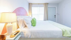 A hotel room with a bed, a bedside table with a lamp, a remote control, and a wall-mounted light fixture. - Charming Beachside Studio: King Bed, AC & Full Kitchen (Fort Lauderdale)