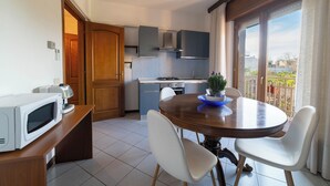Cocina compacta con microondas, estufa y armarios. Se aprecia una mesa de comedor con sillas y una planta en maceta.