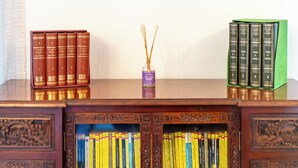 Un escritorio de madera con dos juegos de libros, uno rojo y otro verde, y un pequeño florero morado con ramas de juncos.