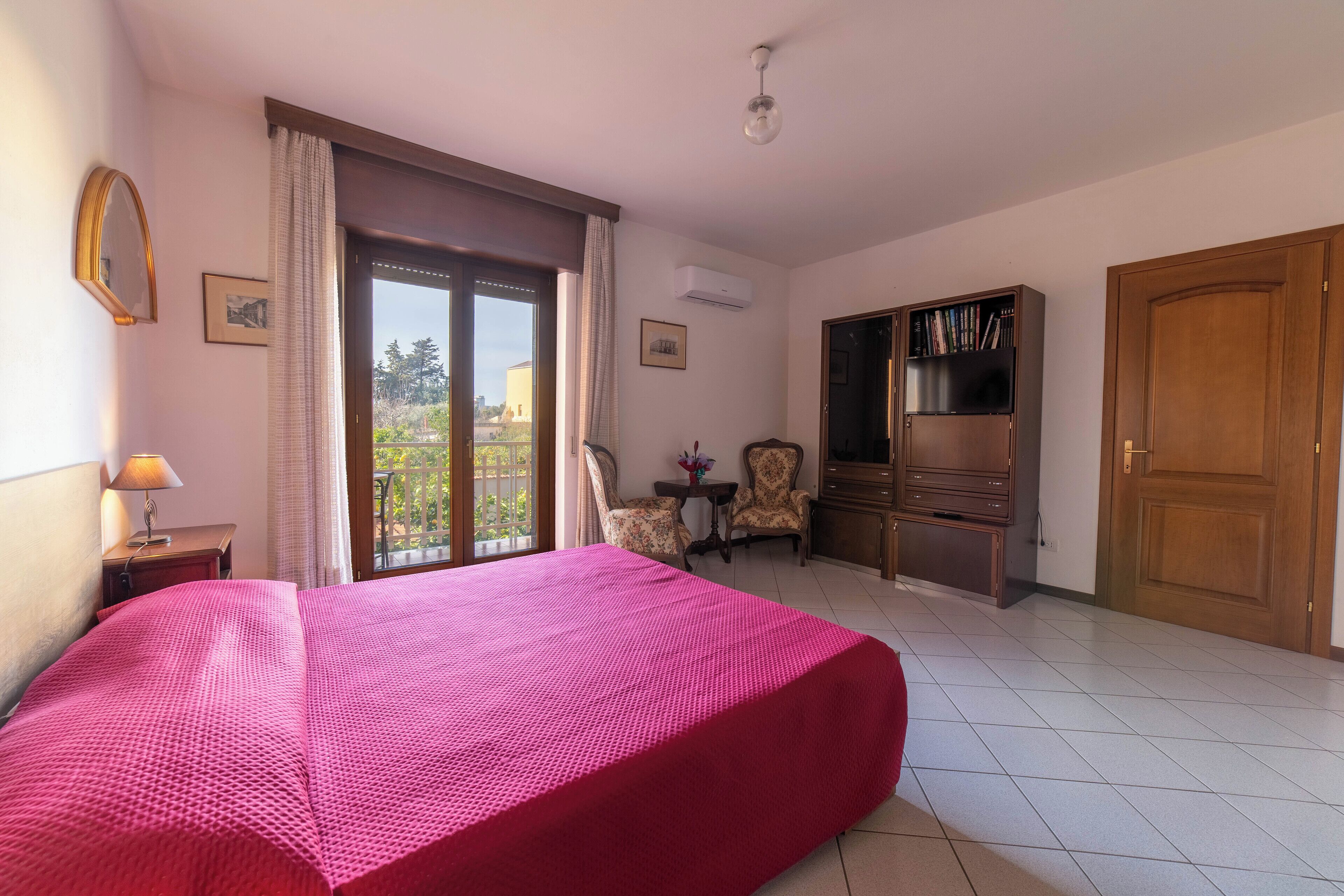 Quarto de hotel com uma cama grande, uma escrivaninha com luminária, uma cadeira, uma mesinha com flores, uma estante e um guarda-roupa.
