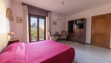 Quarto de hotel com uma cama grande, uma escrivaninha com luminária, uma cadeira, uma mesinha com flores, uma estante e um guarda-roupa.