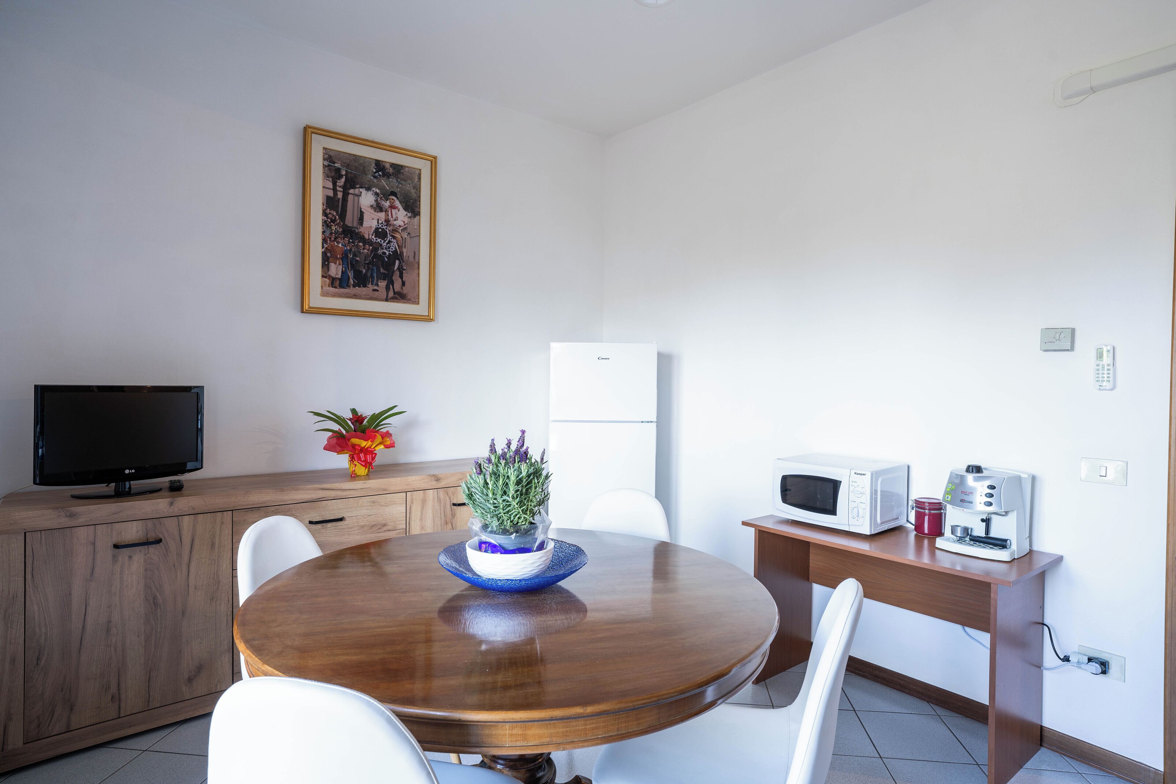 A modern kitchen with a wooden table, white chairs, a television, a microwave, a coffee machine, and a potted plant.