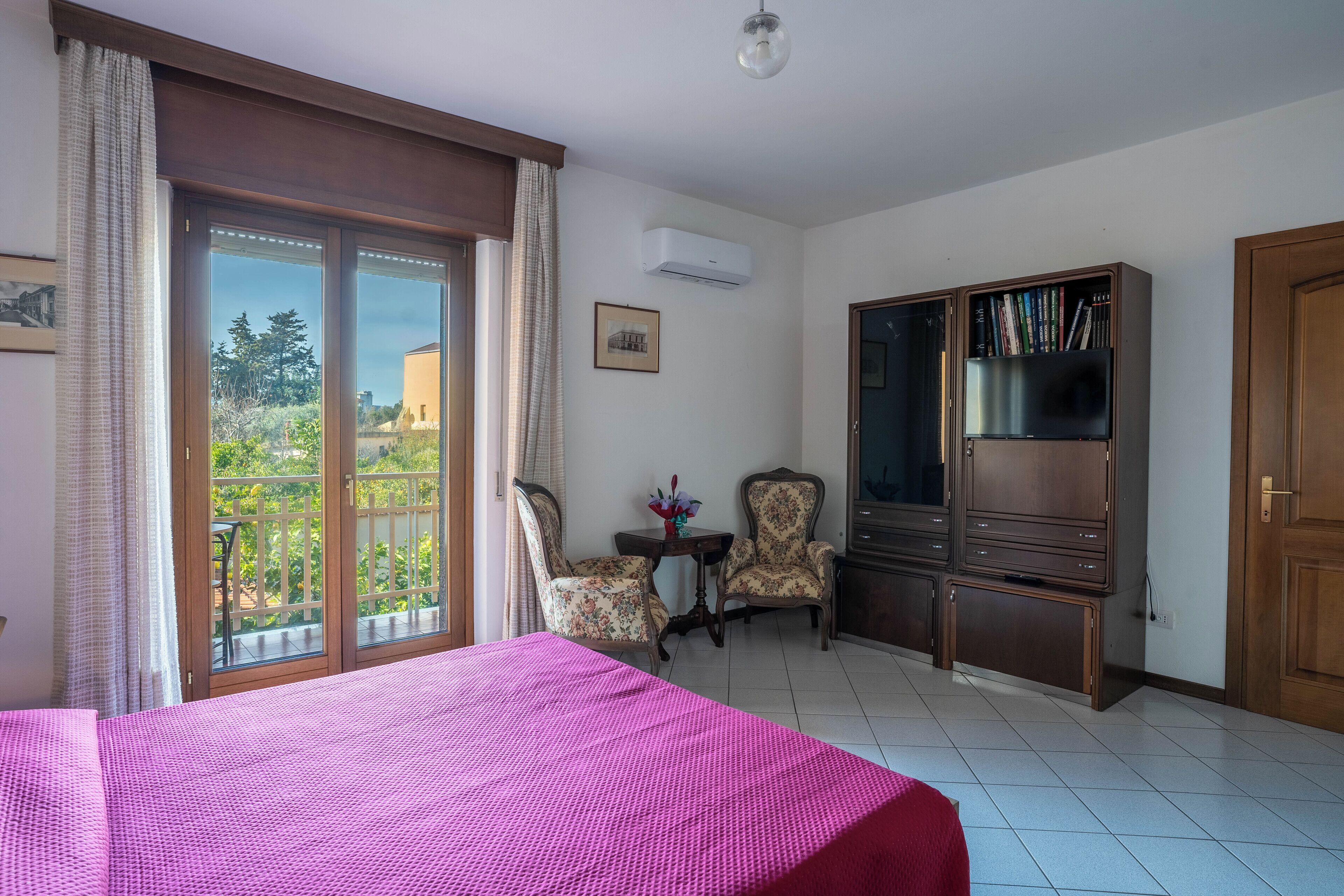 Une chambre d’hôtel comprenant un lit, un bureau, deux chaises, une bibliothèque et une porte-fenêtre coulissante donnant sur l’extérieur.
