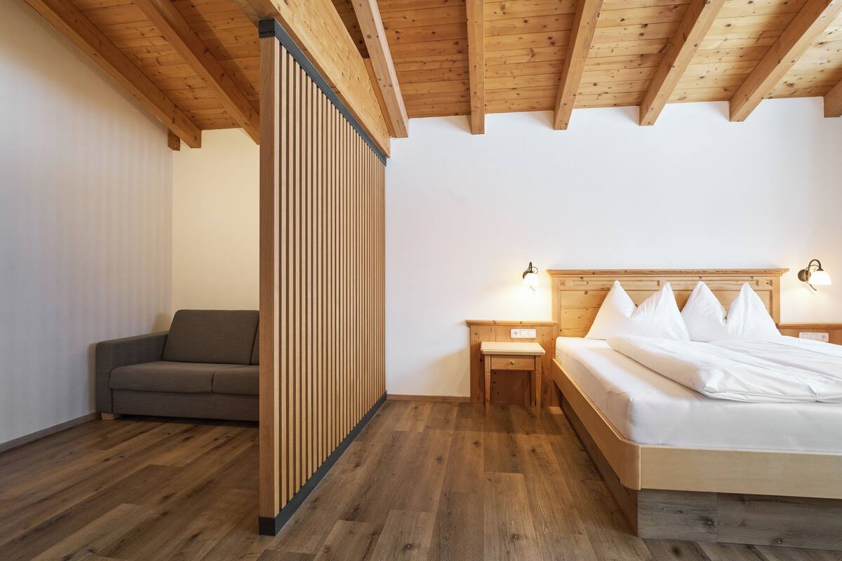 A bedroom with a wooden ceiling, a bed with white bedding, a nightstand with a lamp, and a grey sofa.