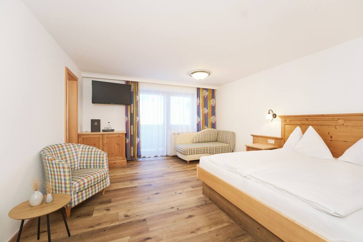 A hotel room with a wooden floor, a bed, a sofa, a chair, a small table, and a TV on the wall.