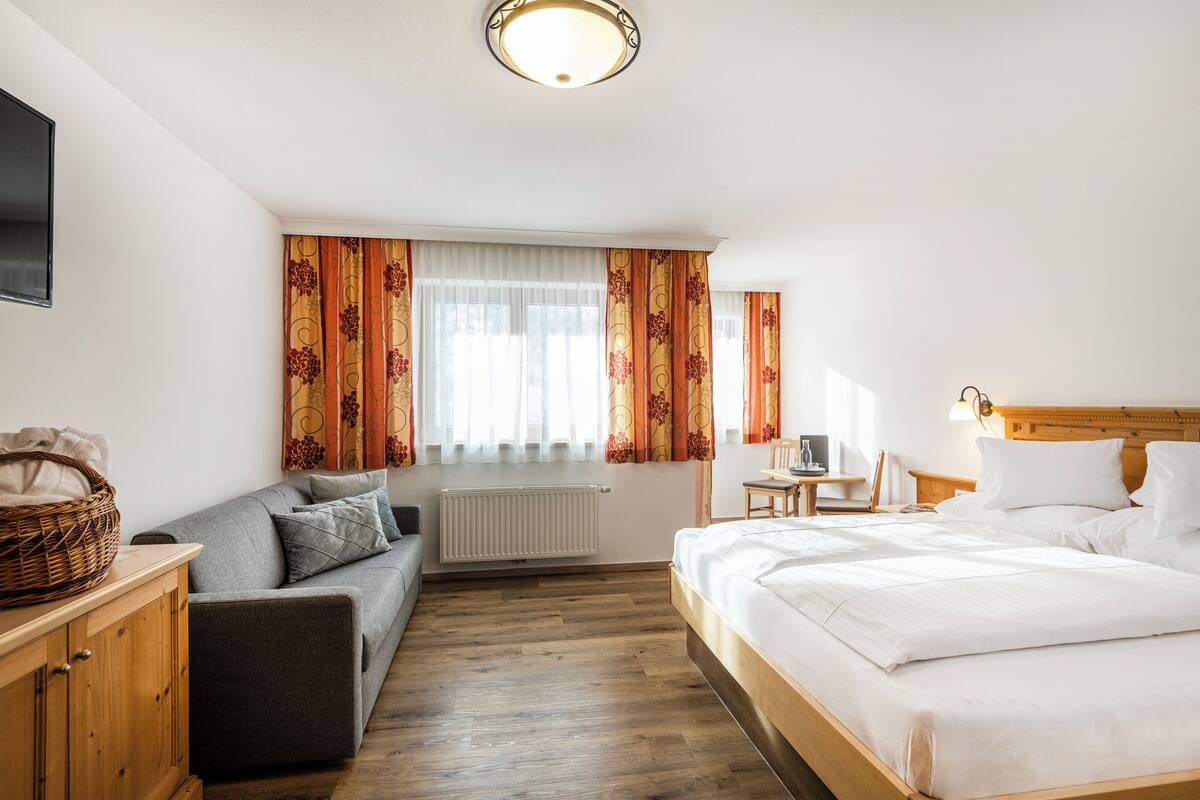 A hotel room with a wooden floor, a grey sofa, a wooden bed, a small table, and a wall-mounted TV.