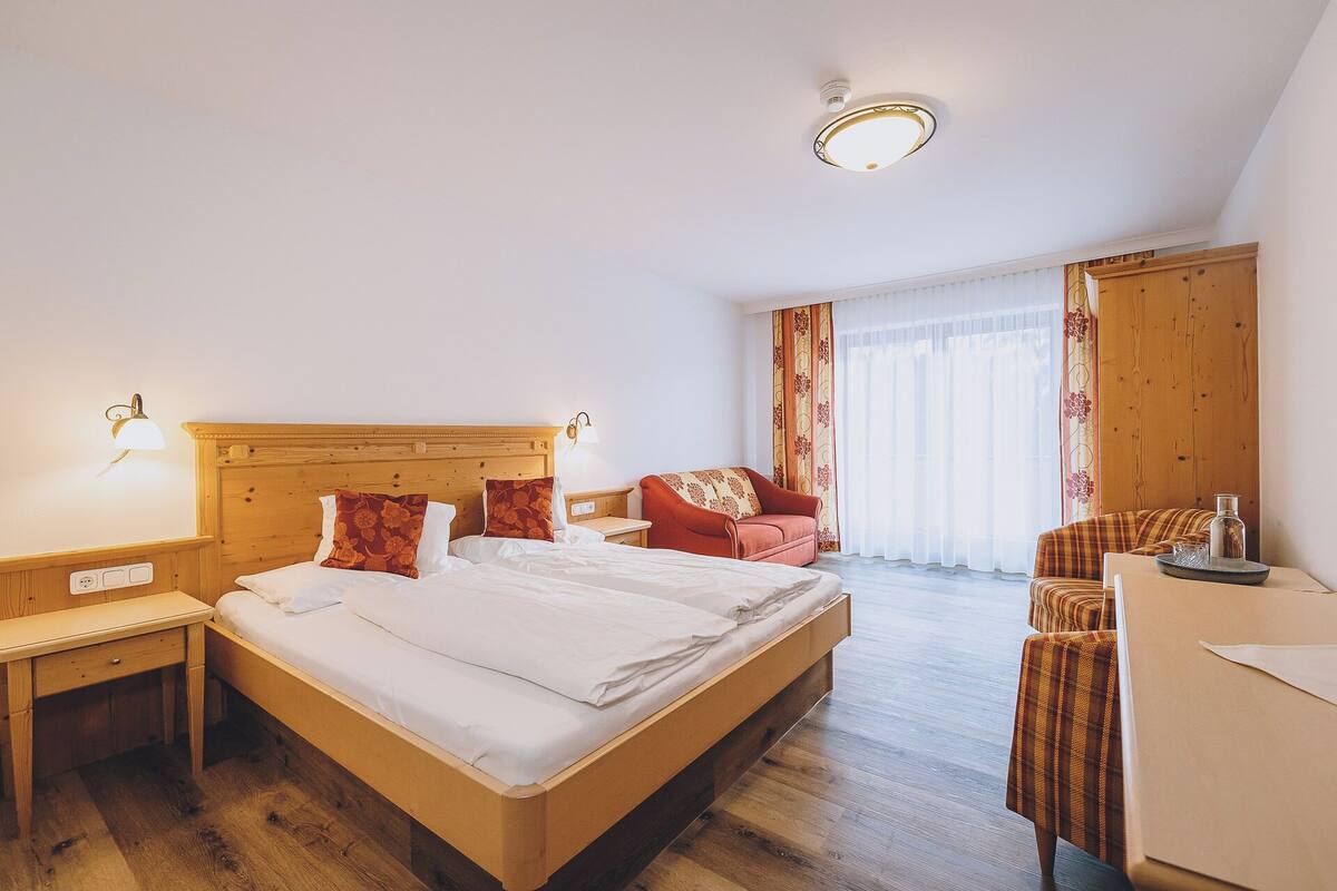 A hotel room with a wooden bed, a desk, a chair, and a large window with curtains.