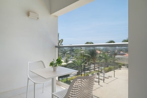 Un balcon avec une table et des chaises blanches, un pot de fleurs et une vue sur un espace aménagé avec des palmiers et un bâtiment situé au loin.
