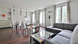 Una sala de estar moderna con un sofá gris, una mesa de centro con libros, una zona de comedor con sillas blancas y ventanas amplias con cortinas.
