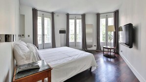 Habitación de hotel con una cama grande, una mesita de noche, un televisor montado en la pared y una ventana con cortinas.