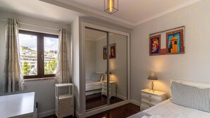 Un dormitorio con cama, mesita de noche, lámpara, espejo y vista a la ciudad por una ventana.