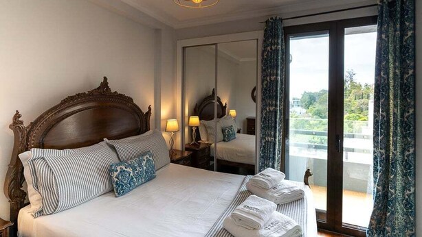 Habitación de hotel con una cama grande, cabecera de madera, dos mesitas de noche con lámparas, un espejo y vistas a través de un ventanal corredero.