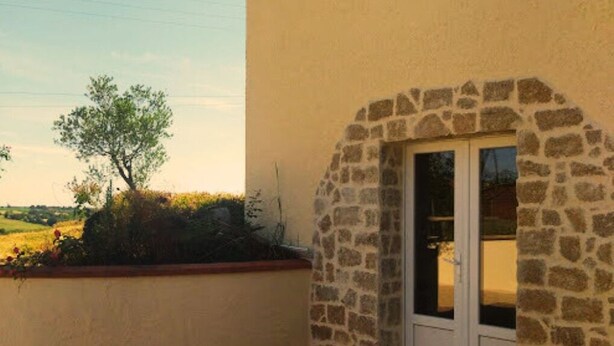 Un bâtiment doté d’une arche en pierre menant à une porte, le tout entouré d’un mur bas et d’un jardin.